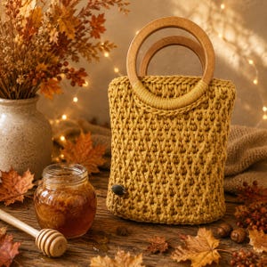 Puede incluir: Un bolso de ganchillo color amarillo mostaza con un patrón de celosía texturizado y dos asas redondas de madera. El bolso tiene una pequeña etiqueta ovalada oscura en la parte inferior izquierda. Está hecho de hilo grueso con tejido uniforme.