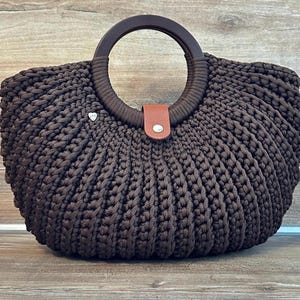 Puede incluir: Un bolso de mano de ganchillo en color marrón oscuro con una silueta redondeada y un patrón de canalé texturizado. El bolso incluye un asa circular de color marrón oscuro y una pequeña solapa de cierre tipo cuero beige con un broche plateado.