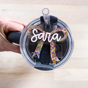 May include: A clear tumbler with a black lid and a straw. The lid has a colorful glitter bow with the name "Sara" written in white.