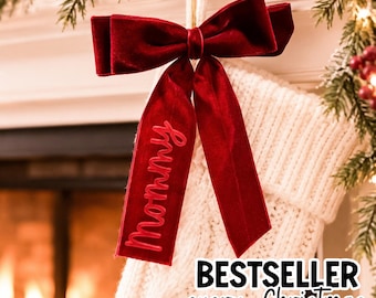 May include: A white cable-knit Christmas stocking featuring a large, deep red velvet bow attached to the top. One of the long velvet ribbon tails is embroidered with the word Mommy in a matching red script font.