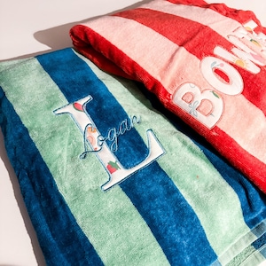 May include: Two striped beach towels. One is red and white, with the name "Bowie" embroidered on it. The other is blue and green, with the name "Logan" embroidered on it. The letter "L" is decorated with a floral pattern.