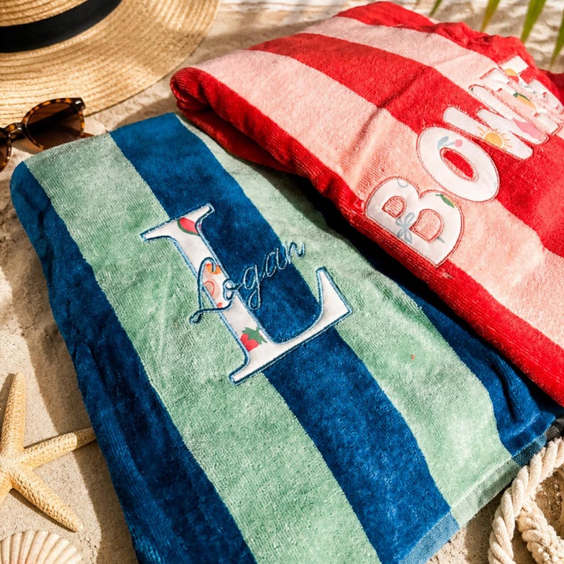 May include: Two beach towels on sand. One is blue and green striped with an embroidered "L" and the name "Logan". The other is red and pink striped with the word "BONNIE". A straw hat and sunglasses are in the background.