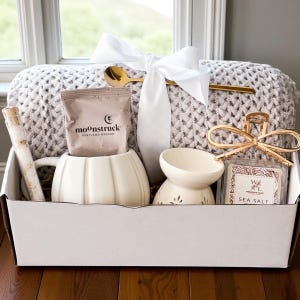 May include: A gift box filled with self-care items. It includes a gray knit blanket, a white pumpkin-shaped mug, a sachet of "moonstruck" tea, a small oil diffuser, a sea salt bar, a gold spoon, and a decorative hair clip. The box is tied with a white ribbon.