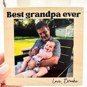 May include: A wooden photo block with the text "Best grandpa ever" in black. The photo shows a man holding a baby. The baby is wearing a pink dress. The text "Love, Brooke" is written in cursive at the bottom.