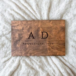 May include: A wooden sign with the initials "A|D" and the date "AUGUST 12TH, 2023" engraved in black.