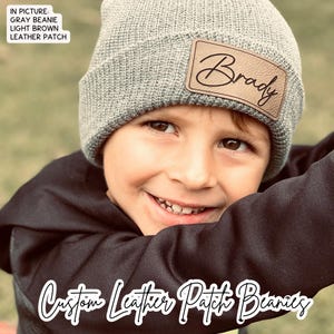 May include: A gray beanie with a light brown leather patch that says "Brady" on it. The beanie is worn by a young boy with a big smile. The text "Custom Leather Patch Beanies" is written in white on a black background.