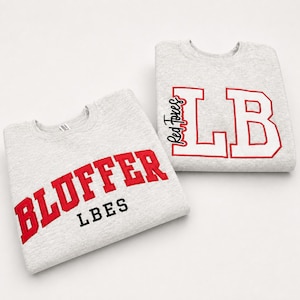 May include: Two light gray crewneck sweatshirts. One sweatshirt features the red text BLUFFER and black text LBES. The other sweatshirt displays the white text LB with a red outline and the red text Red Foxes written vertically along the side.