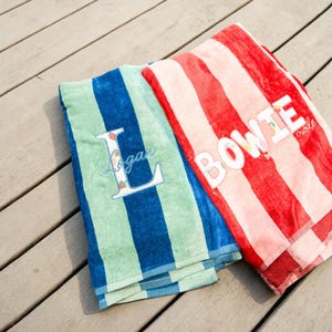 May include: Two striped beach towels. One is blue and green with the name "Logan" embroidered on it. The other is red and pink with the name "Bowie" embroidered on it. Both towels are folded and lying on a wooden surface.