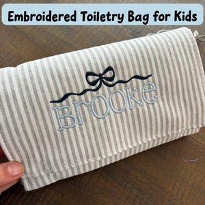 May include: Embroidered toiletry bag for kids with a gray and white striped pattern. The bag features the name "Brooke" embroidered in light blue, with a dark blue bow and wavy line design above the name.