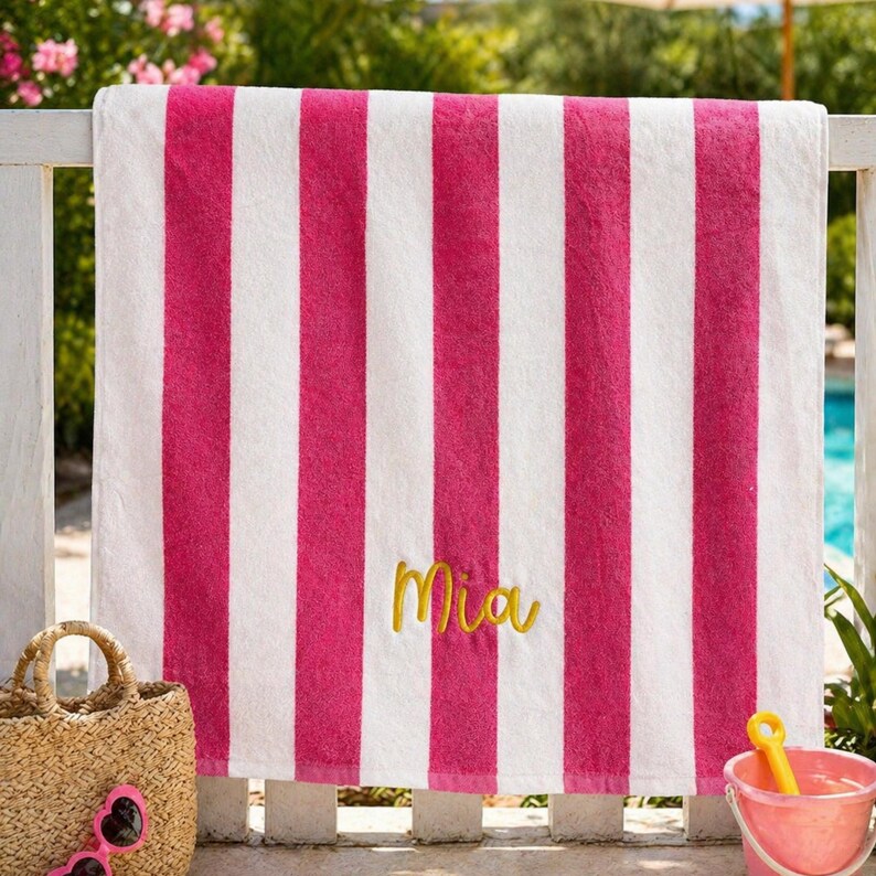 May include: A pink and white striped beach towel with the name "Mia" embroidered in gold script. The towel is hanging on a white fence, with a straw tote bag and pink sunglasses in the foreground.