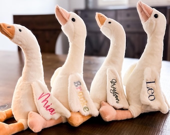 May include: Four plush goose toys in a row, each with a different name embroidered on the wing: Mia, Brooke, Grayson, and Leo. The geese are cream-colored with peach beaks and feet, and are sitting on a wooden surface.
