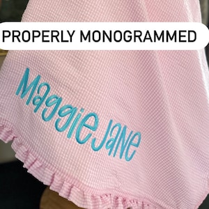 May include: A pink gingham baby blanket with a blue embroidered name "Maggie Jane" on it. The blanket has a ruffled edge.