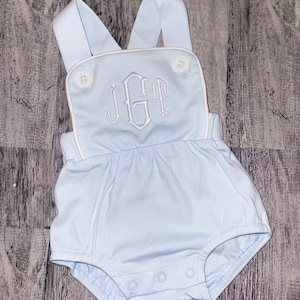 May include: Light blue baby romper with white straps and a white embroidered monogram "JGT" on the front.