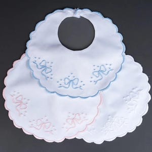 May include: Three embroidered baby bibs with scalloped edges. The top bib is light blue with blue bows, the middle bib is pink with pink bows, and the bottom bib is white with white bows.