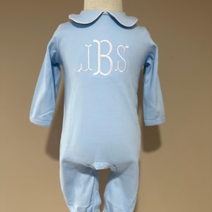 May include: A light blue baby romper with long sleeves and a Peter Pan collar trimmed in white. The romper features a white embroidered monogram on the chest. The garment is displayed on a wooden mannequin.