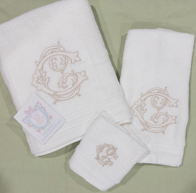 May include: A set of three white, monogrammed bath towels. Each towel features a beige embroidered letter "G" in an ornate, floral design. A small card with a floral crest and the text "PROPERLY MONOGRAMMED" is included.