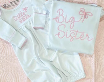 May include: A white long-sleeve infant gown with pink embroidered text reading little sister and a matching white t-shirt with pink embroidered text reading Big Sister. Both garments feature pink stitched bow accents and decorative trim.