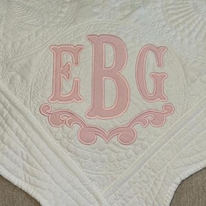 May include: White quilted blanket with a pink embroidered monogram. The monogram features the letters EBG in a decorative font, surrounded by a floral design. The blanket has a detailed, textured pattern.