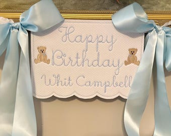 May include: A light blue and white banner with the words "Happy Birthday Whit Campbell" embroidered in light blue script. Two small teddy bear appliqués flank the text. The banner is adorned with large light blue satin bows.