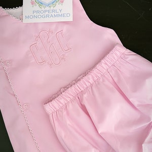 Puede incluir: Conjunto de ropa de beb&eacute; rosa claro, que incluye una parte superior con botones y un pantal&oacute;n corto. La parte superior presenta un monograma bordado y un ribete de encaje blanco. Tambi&eacute;n hay una tarjeta con el texto "PROPERLY MONOGRAMMED".