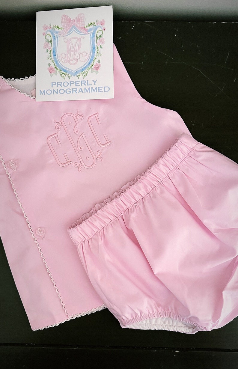 Puede incluir: Conjunto de ropa de beb&eacute; rosa claro, que incluye una parte superior con botones y un pantal&oacute;n corto. La parte superior presenta un monograma bordado y un ribete de encaje blanco. Tambi&eacute;n hay una tarjeta con el texto "PROPERLY MONOGRAMMED".