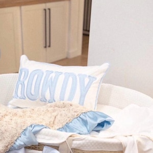 May include: A white pillow with the name "ROWDY" embroidered in light blue, resting in a white bassinet. The bassinet is lined with a soft, cream-colored blanket and a blue satin trim.