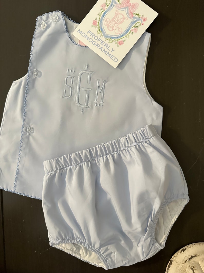 Puede incluir: Conjunto de ropa de beb&eacute; azul claro, que incluye una parte superior con iniciales bordadas y bragas a juego. La parte superior tiene cierres de bot&oacute;n y un borde festoneado. La etiqueta dice "PROPERLY MONOGRAMMED".
