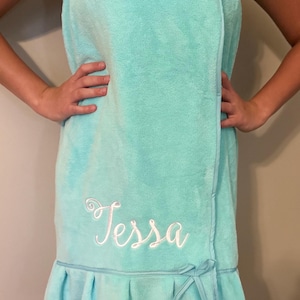 May include: A light turquoise spa wrap towel with the name "Tessa" embroidered in white script. The towel has a ruffled hem and a small bow detail. The wrap is secured with a strap.