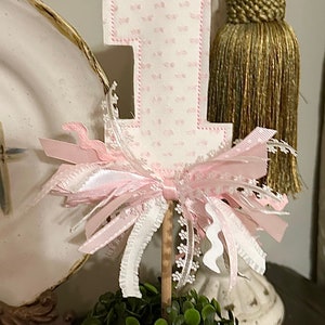 May include: A pink felt number one with a pink and white ribbon bow, attached to a wooden stick. The number one is decorated with a pink and white polka dot pattern. The number one is sitting on top of a small green bush.