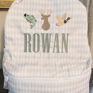 May include: A beige and white checkered backpack with a white zipper and the name "ROWAN" embroidered in green. The backpack features embroidered images of a fish, deer, and duck.