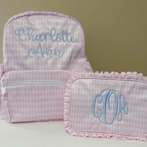 May include: A pink and white gingham backpack and matching cosmetic bag. The backpack has the name "Charlotte Ann" embroidered in light blue. The cosmetic bag has a light blue monogram. Both items have a ruffle trim.