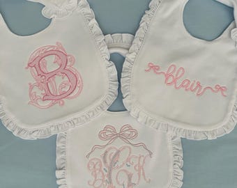 May include: Three white baby bibs with pink embroidered designs. The first bib has a large pink letter "B" with decorative flourishes. The second bib has the name "Blair" embroidered in pink. The third bib has a pink bow and the letters "BCK" embroidered in pink.