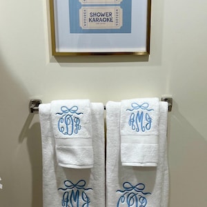May include: White bath towels with blue embroidered monograms and a matching framed print of "Shower Karaoke" tickets. The towels are folded and hanging on a silver towel rack, creating a coordinated bathroom set.