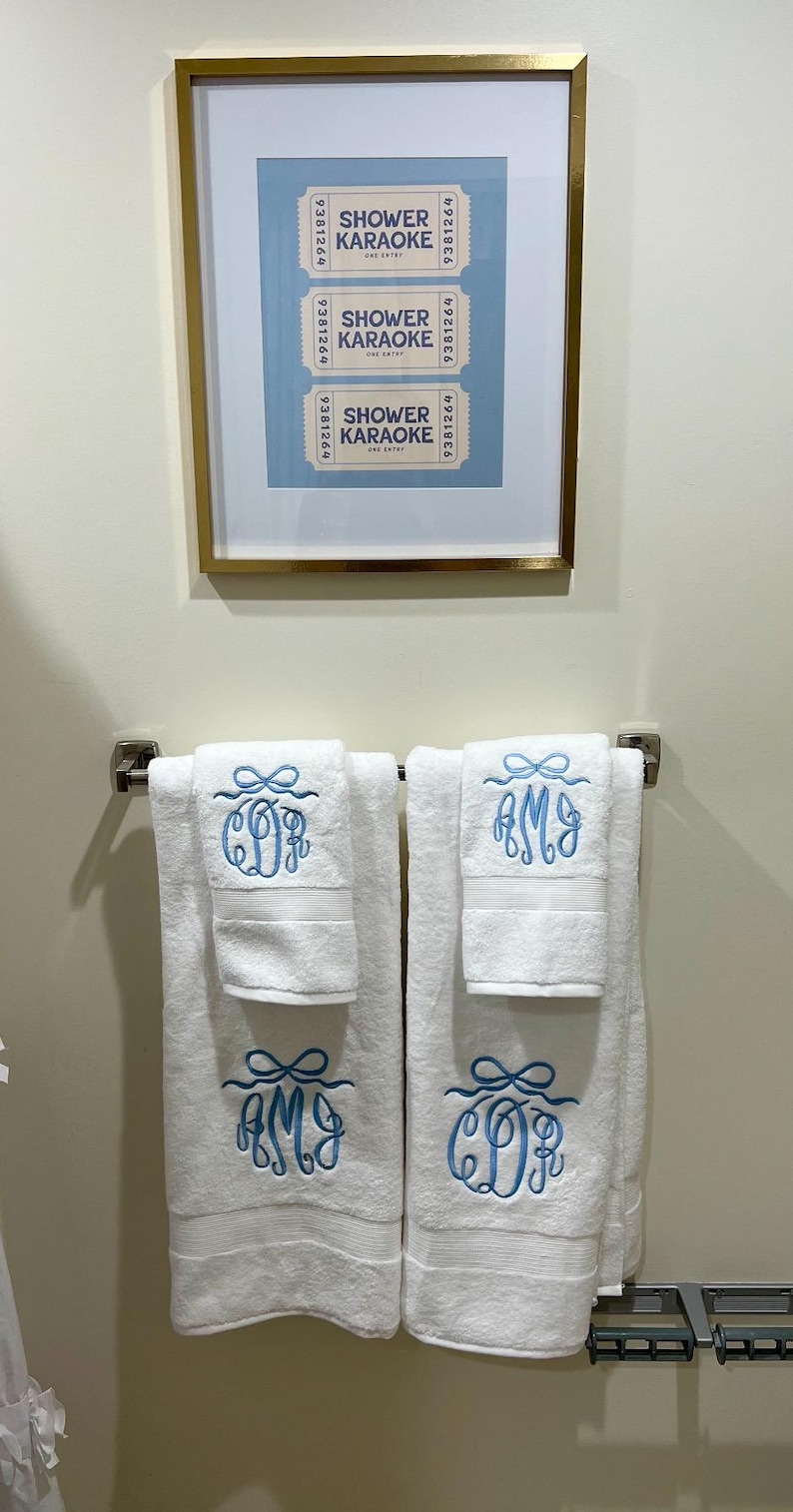 May include: White bath towels with blue embroidered monograms and a matching framed print of "Shower Karaoke" tickets. The towels are folded and hanging on a silver towel rack, creating a coordinated bathroom set.