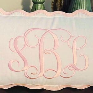May include: A rectangular white pillow with a pink scalloped edge and a large embroidered monogram in light pink. The monogram features the letters SBL in a flowing, cursive style, centered on the pillow.