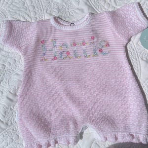 May include: Pink and white striped baby romper with the name "Hattie" embroidered in floral letters. The romper has short sleeves, a ruffled hem, and a white trim around the neckline and sleeves. The fabric has a textured waffle weave.