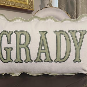 May include: White rectangular pillow with the name "GRADY" embroidered in green. The pillow has a scalloped edge in a lighter green color, adding a decorative touch. The pillow is placed on a dark surface.