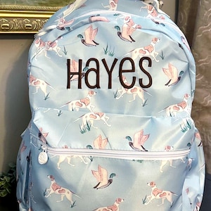 May include: A light blue backpack featuring a repeating pattern of white and brown spotted dogs and flying ducks in green grass. The name Hayes is embroidered in dark brown thread across the center. The bag has a white top handle and white zippers.