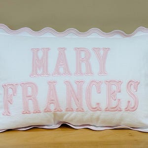 May include: White rectangular pillow with pink scalloped trim and embroidered text reading "MARY FRANCES" in a decorative font. The pillow is on a wooden surface.