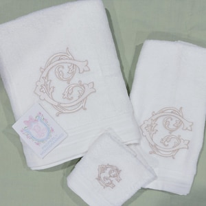 May include: A set of three white monogrammed towels. Each towel features a large, ornate letter "G" embroidered in a light beige color. A small card with a floral crest is visible. The towels are arranged on a light green surface.