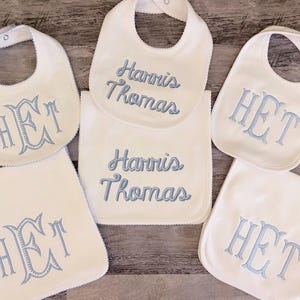 May include: White baby bibs and burp cloths with light blue embroidered lettering. Some items display the initials "HET" in a decorative font, while others feature the name "Harris Thomas". The bibs have a scalloped edge.
