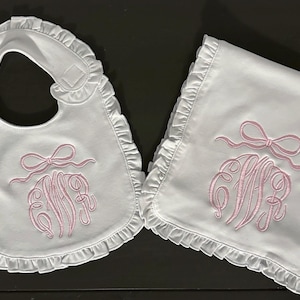 May include: A white baby bib and burp cloth with pink embroidered ruffles and a monogram with a bow.