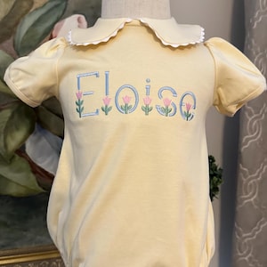 May include: A light yellow baby romper with a Peter Pan collar and short sleeves. The name "Eloise" is embroidered in blue with pink flower accents. The romper is displayed on a mannequin.