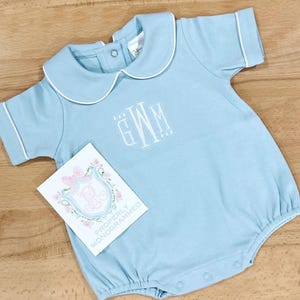 May include: A light blue baby onesie with a white peter pan collar and white piping. The onesie has a white embroidered monogram "GWM" on the front. The onesie is on a wooden surface.