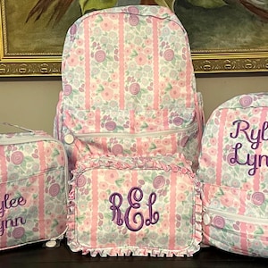 May include: A matching set of floral print backpacks and a lunch bag. The set includes a large backpack, a smaller backpack, a lunch bag, and a small pouch. Each item has a pink, white, and lavender floral pattern and is personalized with a name.