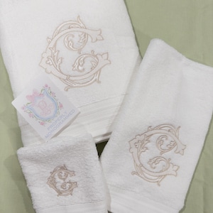 May include: A set of white cotton towels, including a bath towel, hand towel, and washcloth. Each towel is embroidered with a beige, ornate letter "G". A small card with a floral crest and the text "Properly Monogrammed" is also present.