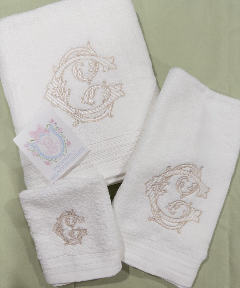 May include: A set of white cotton towels, including a bath towel, hand towel, and washcloth. Each towel is embroidered with a beige, ornate letter "G". A small card with a floral crest and the text "Properly Monogrammed" is also present.