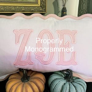 May include: White rectangular pillow with a pink scalloped edge and the name "ZOE" embroidered in pink. The text "Properly Monogrammed" is below the name. Two pumpkins are in the foreground.