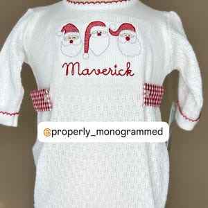 May include: White knitted baby onesie with red trim and three embroidered Santa faces. The onesie has a red and white gingham ribbon around the waist and the name "Maverick" embroidered below the Santa faces.