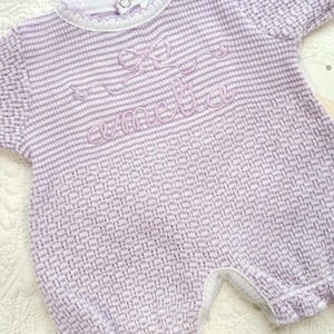 May include: A light purple baby romper with horizontal stripes and the name "Amelia" embroidered in cursive. The romper has short sleeves, a round neckline, and a decorative bow above the name. The fabric has a textured appearance.